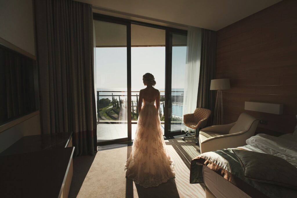 Bride opening window balcony in soft light in hotel room.